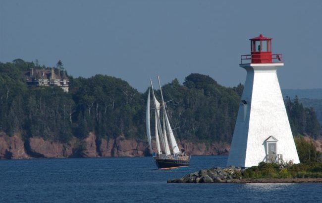 Touring sail boat passes the lighthouse at Baddeck on the Bras d'Or Lake. In the background is the former summer home of Alexander Graham Bell.