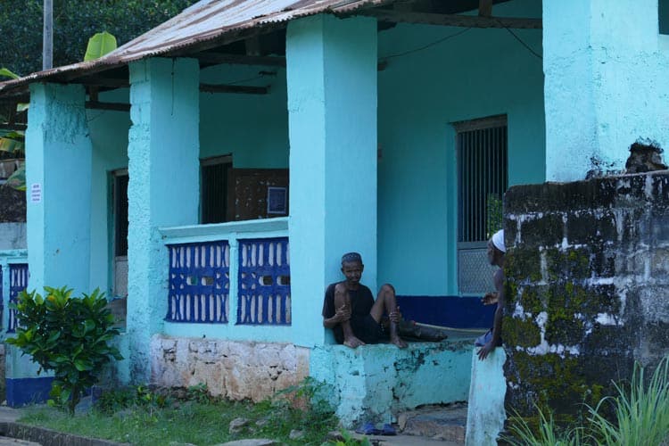 >A man sitting by the bright blue cottage in the village of Maradoka. Photo by Adrian Rorvik