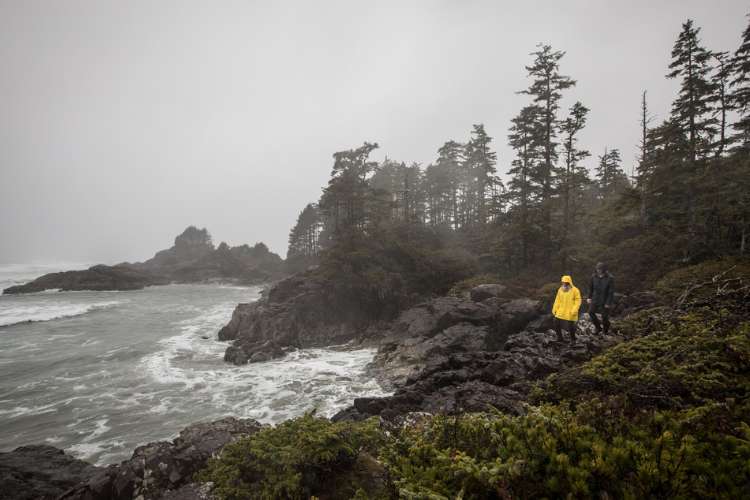 A person in a bright yellow slicker walks on the rocks facing the ocean during a storm.