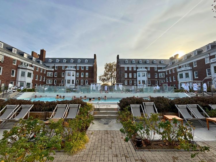 Empty deck chairs sit in front of a large steamy pool where a few people are soaking.