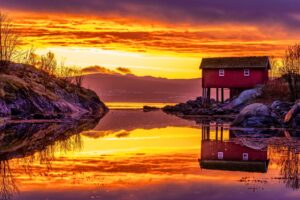 The fiery golds and oranges of Norway's midnight sun illuminate a classic Norwegian boathouse. Photo by Anders Vestin from Getty Images via Canva