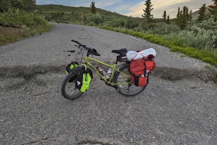 A bicycle shown near a one-foot drop on a closed road, marked by rocks in an X-shape and a red flag
