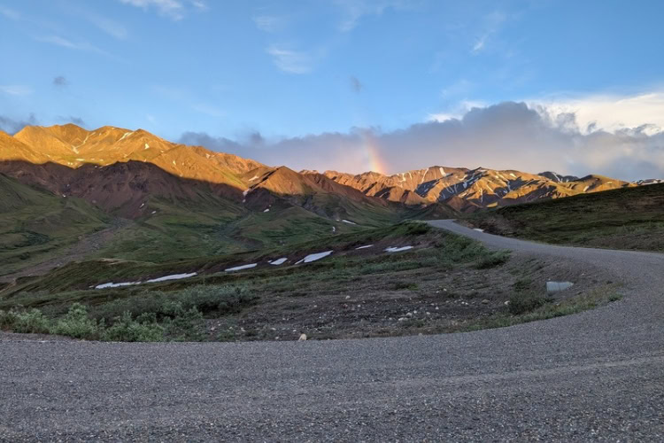 The gravel road beyond the Pretty Rocks landslide, with sunlit mountain tops and a rainbow in the clouds during the late evening.