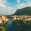 Brown-arched bridge surrounded by stone buildings and leafy green trees in Mostar, Bosnia and Herzegovina. Photo by Yu Siang Teo, Unsplash