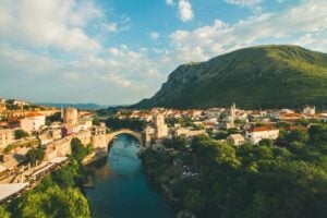 Brown-arched bridge surrounded by stone buildings and leafy green trees in Mostar, Bosnia and Herzegovina. Photo by Yu Siang Teo, Unsplash