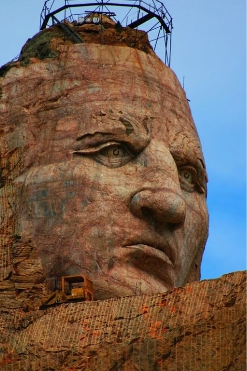 The 87-foot tall face of Crazy Horse at the Crazy Horse Memorial in South Dakota, a full one-third taller than any face on Mount Rushmore, dwarfs a yellow crawler tractor in 2010. Photo by Frank Hosek